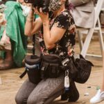 Snohomish Wedding Photographer GSquared Weddings A photographer wearing a floral shirt, gray pants, and a patterned face mask kneels on the ground, holding a camera at a seated event. She has camera bags at her waist and is surrounded by scattered flower petals. Serving Seattle, Snohomish and Orlando