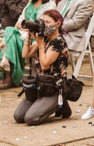 Snohomish Wedding Photographer GSquared Weddings A photographer wearing a floral shirt, gray pants, and a patterned face mask kneels on the ground, holding a camera at a seated event. She has camera bags at her waist and is surrounded by scattered flower petals. Serving Seattle, Snohomish and Orlando
