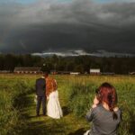 Snohomish Wedding Photographer GSquared Weddings A photographer captures a bride and groom standing together on a grassy path, facing away toward a dramatic cloudy sky and distant barns, surrounded by green fields. Serving Seattle, Snohomish and Orlando