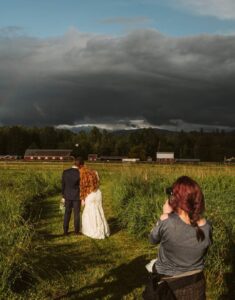 Snohomish Wedding Photographer GSquared Weddings A photographer captures a bride and groom standing together on a grassy path, facing away toward a dramatic cloudy sky and distant barns, surrounded by green fields. Serving Seattle, Snohomish and Orlando
