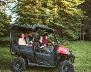 Snohomish Wedding Photographer GSquared Weddings Four people sit and smile in a black and red off-road ATV parked on grass, with tall green pine trees in the background. Sunlight filters through the trees, creating a warm, cheerful atmosphere. Serving Seattle, Snohomish and Orlando