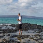 Snohomish Wedding Photographer GSquared Weddings A man stands on rocky shore with a camera, wearing a white shirt and black shorts, looking out at turquoise ocean waves. Distant hills and a partly cloudy sky are visible in the background. Serving Seattle, Snohomish and Orlando
