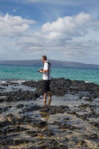 Snohomish Wedding Photographer GSquared Weddings A man stands on rocky shore with a camera, wearing a white shirt and black shorts, looking out at turquoise ocean waves. Distant hills and a partly cloudy sky are visible in the background. Serving Seattle, Snohomish and Orlando