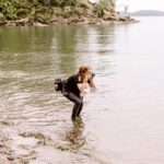 Snohomish Wedding Photographer GSquared Weddings A person wearing black clothes and a camera bag stands knee-deep in a calm lake, photographing something onshore. Trees and rocky land appear in the background under an overcast sky. Serving Seattle, Snohomish and Orlando