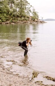 Snohomish Wedding Photographer GSquared Weddings A person wearing black clothes and a camera bag stands knee-deep in a calm lake, photographing something onshore. Trees and rocky land appear in the background under an overcast sky. Serving Seattle, Snohomish and Orlando