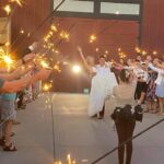 Snohomish Wedding Photographer GSquared Weddings A bride and groom walk joyfully through a path of guests holding sparklers, with the groom carrying the bride in his arms outside a building. A photographer captures the moment. Serving Seattle, Snohomish and Orlando