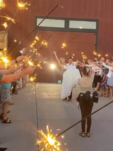 Snohomish Wedding Photographer GSquared Weddings A bride and groom walk joyfully through a path of guests holding sparklers, with the groom carrying the bride in his arms outside a building. A photographer captures the moment. Serving Seattle, Snohomish and Orlando
