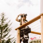 Snohomish Wedding Photographer GSquared Weddings A woman with long hair stands on a wooden deck, holding a camera up to her face. She is wearing a floral dress and has a camera bag at her side. There is a potted plant with flowers next to her and a tree in the background. Serving Seattle, Snohomish and Orlando