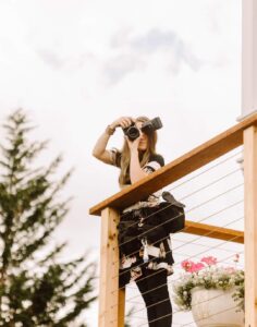 Snohomish Wedding Photographer GSquared Weddings A woman with long hair stands on a wooden deck, holding a camera up to her face. She is wearing a floral dress and has a camera bag at her side. There is a potted plant with flowers next to her and a tree in the background. Serving Seattle, Snohomish and Orlando