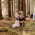 Snohomish Wedding Photographer GSquared Weddings A wedding party stands in a forest of tall trees. The bride in white and bridesmaids in purple dresses pose with groomsmen in gray suits. A photographer adjusts the bride’s dress as sunlight filters through the trees. Serving Seattle, Snohomish and Orlando