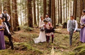 Snohomish Wedding Photographer GSquared Weddings A wedding party stands in a forest of tall trees. The bride in white and bridesmaids in purple dresses pose with groomsmen in gray suits. A photographer adjusts the bride’s dress as sunlight filters through the trees. Serving Seattle, Snohomish and Orlando