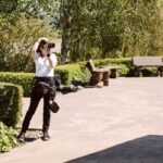 Snohomish Wedding Photographer GSquared Weddings A photographer dressed in black and white stands on a paved area outdoors, taking a photo. They are surrounded by green hedges, trees, benches, and garden decor on a sunny day. Serving Seattle, Snohomish and Orlando