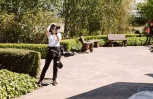Snohomish Wedding Photographer GSquared Weddings A photographer dressed in black and white stands on a paved area outdoors, taking a photo. They are surrounded by green hedges, trees, benches, and garden decor on a sunny day. Serving Seattle, Snohomish and Orlando