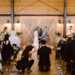 Snohomish Wedding Photographer GSquared Weddings A bride and groom stand at the altar in a rustic barn, holding hands and facing each other as they exchange vows. Wedding guests are seated, some taking photos, with bridesmaids and groomsmen standing nearby. Serving Seattle, Snohomish and Orlando