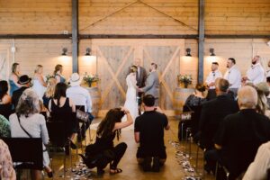 Snohomish Wedding Photographer GSquared Weddings A bride and groom stand at the altar in a rustic barn, holding hands and facing each other as they exchange vows. Wedding guests are seated, some taking photos, with bridesmaids and groomsmen standing nearby. Serving Seattle, Snohomish and Orlando