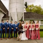 Snohomish Wedding Photographer GSquared Weddings A wedding party poses outdoors, with five men in blue suits and four women in pink dresses standing beside a bride in white. A photographer stands in front, preparing to take their picture. A building and silo are in the background. Serving Seattle, Snohomish and Orlando