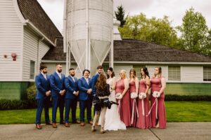 Snohomish Wedding Photographer GSquared Weddings A wedding party poses outdoors, with five men in blue suits and four women in pink dresses standing beside a bride in white. A photographer stands in front, preparing to take their picture. A building and silo are in the background. Serving Seattle, Snohomish and Orlando