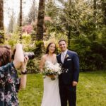 Snohomish Wedding Photographer GSquared Weddings A bride and groom stand together outdoors, smiling as a photographer takes their picture. The bride holds a bouquet of white flowers, and the couple is surrounded by lush green trees and grass. Serving Seattle, Snohomish and Orlando