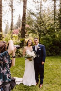 Snohomish Wedding Photographer GSquared Weddings A bride and groom stand together outdoors, smiling as a photographer takes their picture. The bride holds a bouquet of white flowers, and the couple is surrounded by lush green trees and grass. Serving Seattle, Snohomish and Orlando
