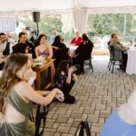 Snohomish Wedding Photographer GSquared Weddings A group of people seated at tables and on the ground under a white tent at an outdoor event, attentively watching something off camera. Trees and a pond are visible in the background. Serving Seattle, Snohomish and Orlando