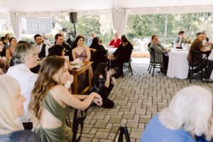 Snohomish Wedding Photographer GSquared Weddings A group of people seated at tables and on the ground under a white tent at an outdoor event, attentively watching something off camera. Trees and a pond are visible in the background. Serving Seattle, Snohomish and Orlando