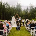 Snohomish Wedding Photographer GSquared Weddings A wedding ceremony takes place outdoors, with guests seated on white chairs facing a couple and officiant under a wooden arch, surrounded by tall trees and greenery. Serving Seattle, Snohomish and Orlando