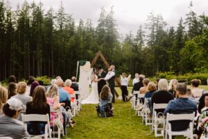 Snohomish Wedding Photographer GSquared Weddings A wedding ceremony takes place outdoors, with guests seated on white chairs facing a couple and officiant under a wooden arch, surrounded by tall trees and greenery. Serving Seattle, Snohomish and Orlando