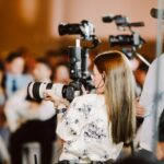 Snohomish Wedding Photographer GSquared Weddings A woman with long brown hair holds a large camera, photographing an event indoors. People sit in the blurred background, and other videographers with equipment are nearby. The setting appears lively and professionally covered. Serving Seattle, Snohomish and Orlando