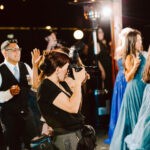 Snohomish Wedding Photographer GSquared Weddings A photographer takes pictures at a lively indoor event, focusing on guests dressed in formal attire, including women and girls in blue dresses, under warm string lights. Serving Seattle, Snohomish and Orlando