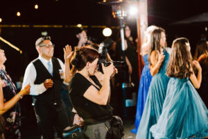 Snohomish Wedding Photographer GSquared Weddings A photographer takes pictures at a lively indoor event, focusing on guests dressed in formal attire, including women and girls in blue dresses, under warm string lights. Serving Seattle, Snohomish and Orlando