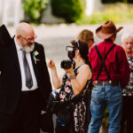 Snohomish Wedding Photographer GSquared Weddings A man in a suit and tie talks with a woman holding a camera at an outdoor event. Other people, including a person in a cowboy hat and suspenders, stand in the background. Serving Seattle, Snohomish and Orlando