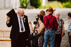 Snohomish Wedding Photographer GSquared Weddings A man in a suit and tie talks with a woman holding a camera at an outdoor event. Other people, including a person in a cowboy hat and suspenders, stand in the background. Serving Seattle, Snohomish and Orlando