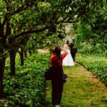 Snohomish Wedding Photographer GSquared Weddings A photographer in a red shirt takes pictures of a bride and groom standing together on a green, leafy pathway surrounded by lush trees and vegetation. Serving Seattle, Snohomish and Orlando
