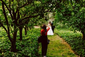 Snohomish Wedding Photographer GSquared Weddings A photographer in a red shirt takes pictures of a bride and groom standing together on a green, leafy pathway surrounded by lush trees and vegetation. Serving Seattle, Snohomish and Orlando