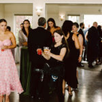Snohomish Wedding Photographer GSquared Weddings Guests mingle at an indoor event. A woman in a pink floral dress holds a drink, while another woman in black carries two drinks and photography gear. More people gather and converse in the background near large windows. Serving Seattle, Snohomish and Orlando