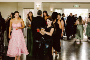 Snohomish Wedding Photographer GSquared Weddings Guests mingle at an indoor event. A woman in a pink floral dress holds a drink, while another woman in black carries two drinks and photography gear. More people gather and converse in the background near large windows. Serving Seattle, Snohomish and Orlando