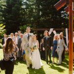 Snohomish Wedding Photographer GSquared Weddings A bride and groom walk arm in arm outdoors, smiling, followed by a joyful group of guests. Sunlight filters through trees, and a photographer captures the moment in a lush garden setting. Serving Seattle, Snohomish and Orlando