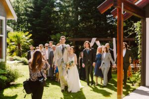 Snohomish Wedding Photographer GSquared Weddings A bride and groom walk arm in arm outdoors, smiling, followed by a joyful group of guests. Sunlight filters through trees, and a photographer captures the moment in a lush garden setting. Serving Seattle, Snohomish and Orlando