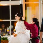 Snohomish Wedding Photographer GSquared Weddings A woman in a white lace wedding dress is being assisted by another woman in a red dress, adjusting her hair. They are indoors, with warm lighting and large windows in the background. Serving Seattle, Snohomish and Orlando