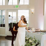 Snohomish Wedding Photographer GSquared Weddings A smiling bride in a white gown stands by a dessert table with cakes and cupcakes, holding a drink. Someone crouches behind her, adjusting her dress. The scene is indoors, with large windows and floral arrangements. Serving Seattle, Snohomish and Orlando