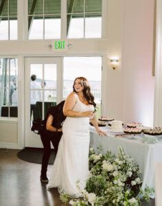 Snohomish Wedding Photographer GSquared Weddings A smiling bride in a white gown stands by a dessert table with cakes and cupcakes, holding a drink. Someone crouches behind her, adjusting her dress. The scene is indoors, with large windows and floral arrangements. Serving Seattle, Snohomish and Orlando