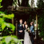 Snohomish Wedding Photographer GSquared Weddings A bride and groom stand under a wooden arbor surrounded by greenery, while a woman holding a bouquet stands beside them, arranging something for the bride. The groom wears a suit and the bride is in a white dress, smiling. Serving Seattle, Snohomish and Orlando