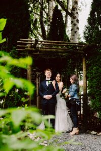 Snohomish Wedding Photographer GSquared Weddings A bride and groom stand under a wooden arbor surrounded by greenery, while a woman holding a bouquet stands beside them, arranging something for the bride. The groom wears a suit and the bride is in a white dress, smiling. Serving Seattle, Snohomish and Orlando