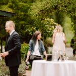 Snohomish Wedding Photographer GSquared Weddings A group of people socialize outdoors near greenery; one woman with a camera smiles while talking, others stand nearby, and a white round table with drinks is in the foreground. Serving Seattle, Snohomish and Orlando