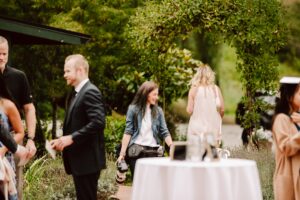 Snohomish Wedding Photographer GSquared Weddings A group of people socialize outdoors near greenery; one woman with a camera smiles while talking, others stand nearby, and a white round table with drinks is in the foreground. Serving Seattle, Snohomish and Orlando