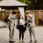 Snohomish Wedding Photographer GSquared Weddings Three people stand outdoors on gravel; two men in grey suits, one wearing a mask, listen to a woman in casual clothes holding papers. A gazebo and wooden fence are in the background. Serving Seattle, Snohomish and Orlando