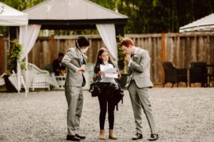 Snohomish Wedding Photographer GSquared Weddings Three people stand outdoors on gravel; two men in grey suits, one wearing a mask, listen to a woman in casual clothes holding papers. A gazebo and wooden fence are in the background. Serving Seattle, Snohomish and Orlando
