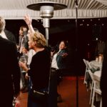 Snohomish Wedding Photographer GSquared Weddings A group of people in semi-formal attire are gathered at an indoor event under a tent, some chatting and holding drinks, while a woman in the center smiles and waves enthusiastically at the camera. Serving Seattle, Snohomish and Orlando