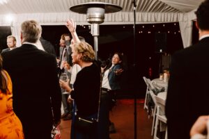 Snohomish Wedding Photographer GSquared Weddings A group of people in semi-formal attire are gathered at an indoor event under a tent, some chatting and holding drinks, while a woman in the center smiles and waves enthusiastically at the camera. Serving Seattle, Snohomish and Orlando