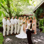 Snohomish Wedding Photographer GSquared Weddings A bride and groom stand with their wedding party, all dressed in light colors, posing for a photo outdoors near a wooden building surrounded by greenery. A photographer faces them, preparing to take the picture. Serving Seattle, Snohomish and Orlando